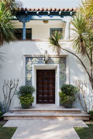 exterior view of front door with tiled surround in traditional spanish colonial style home with white render and palm trees steps to front door