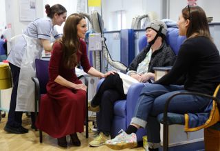 Kate Middleton wearing a red sweater and skirt sitting in a chair and putting her hand on the knee of a woman sitting in a chair wearing a cold cap and getting cancer treatment