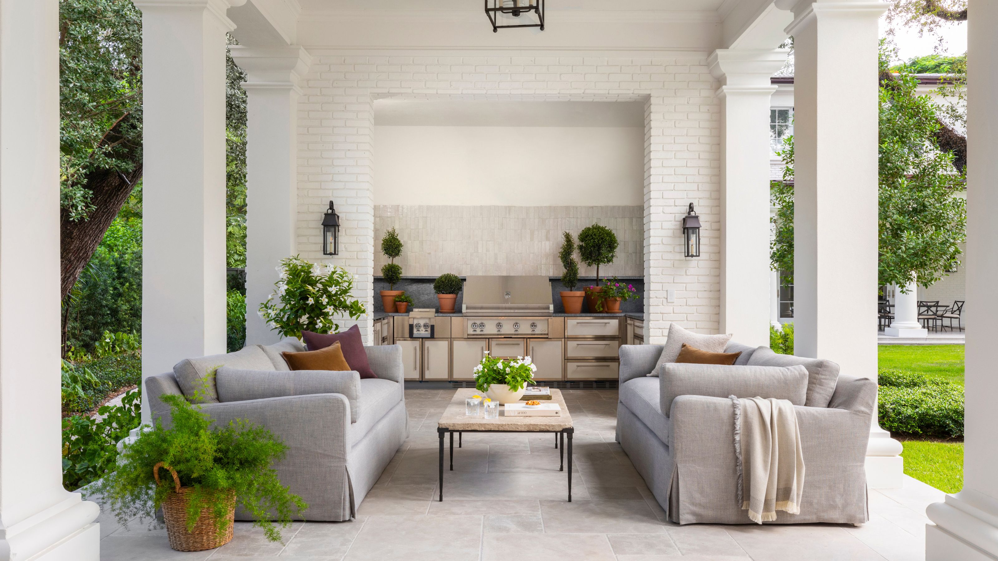 a white brick pillared covered patio area with a large outdoor kitchen, two linen couches, a coffee table and potted plants