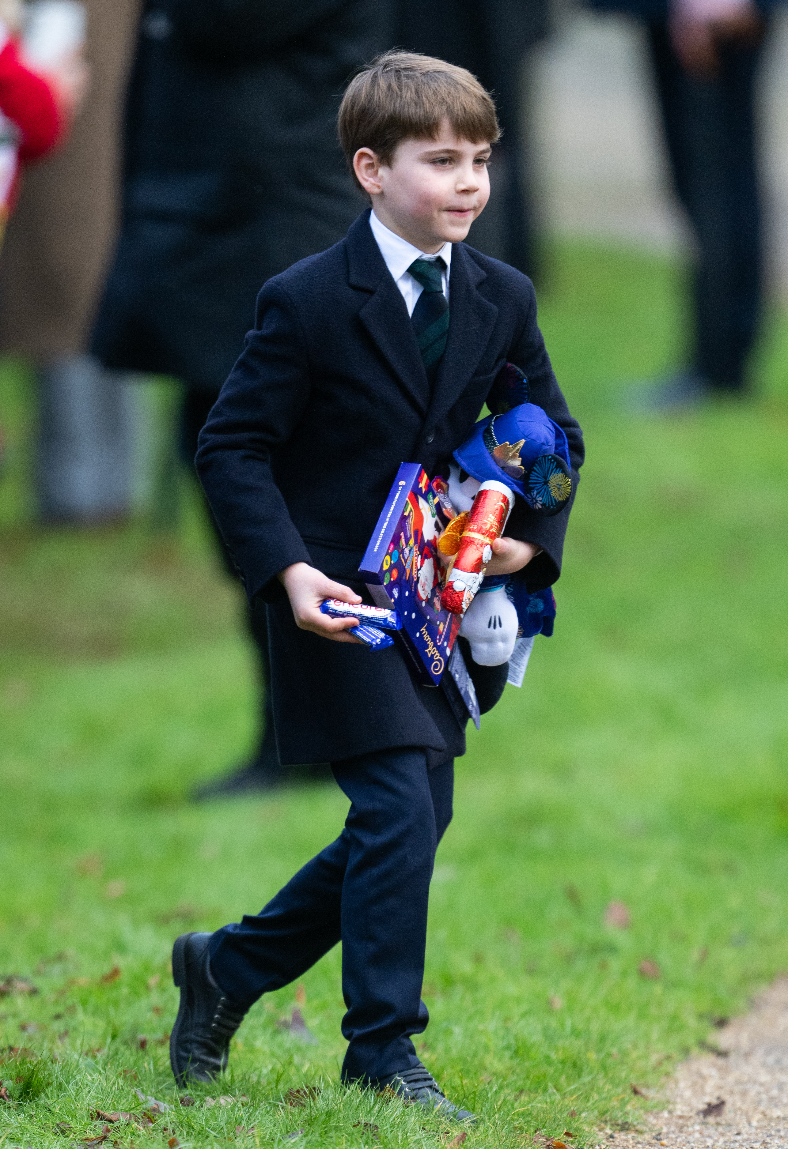 Prince Louis walking in grass wearing a suit and carrying toys