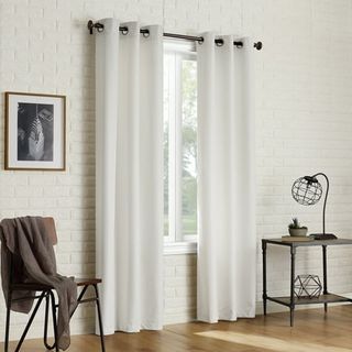 Some white thermal curtains hanging from a metal curtain rod in a white brick room. A chair draped with a blanket and a side table with a small lap on either side.