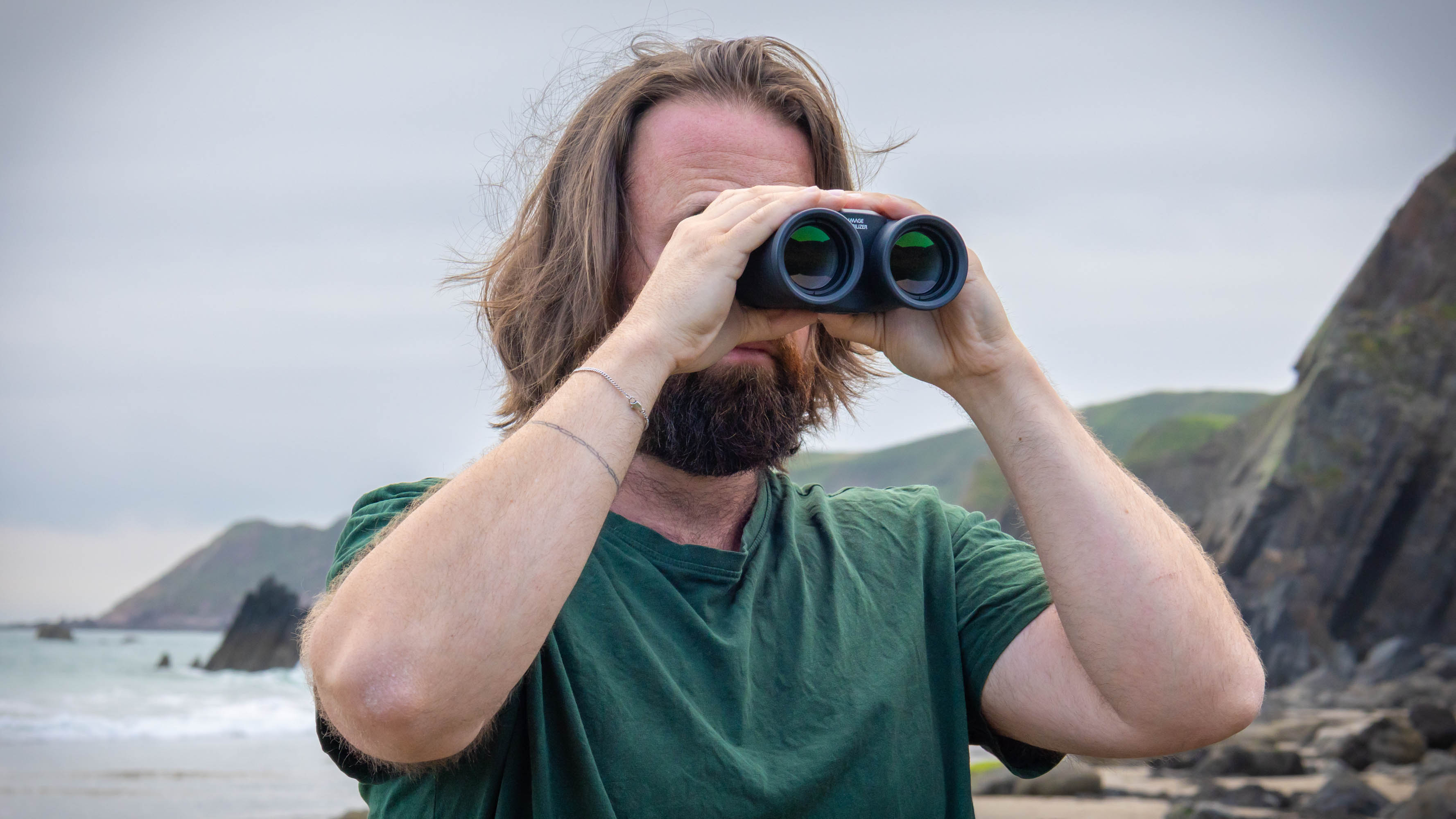 A male using the Canon 12x36 IS III binoculars with the sea and coast behind him.