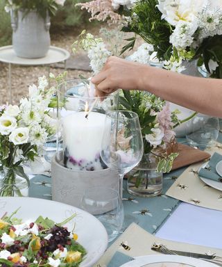 A pillar candle being lit with a maatch on an outdoor table displayed with cut flowers