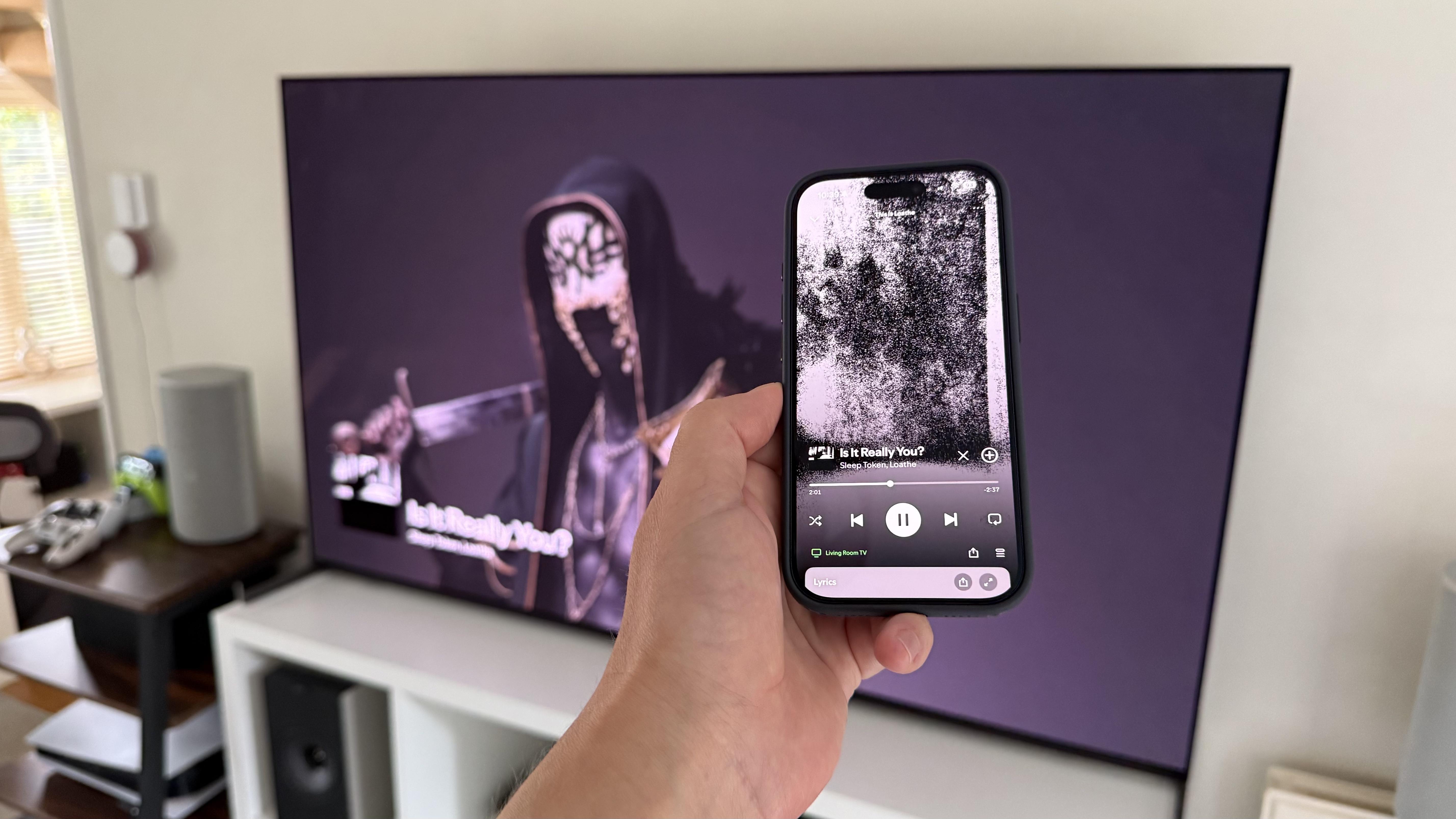 A photo of an iPhone being held in front of a TV. On the phone screen is the Spotify app, and in the background is artwork from the music being played on the TV.