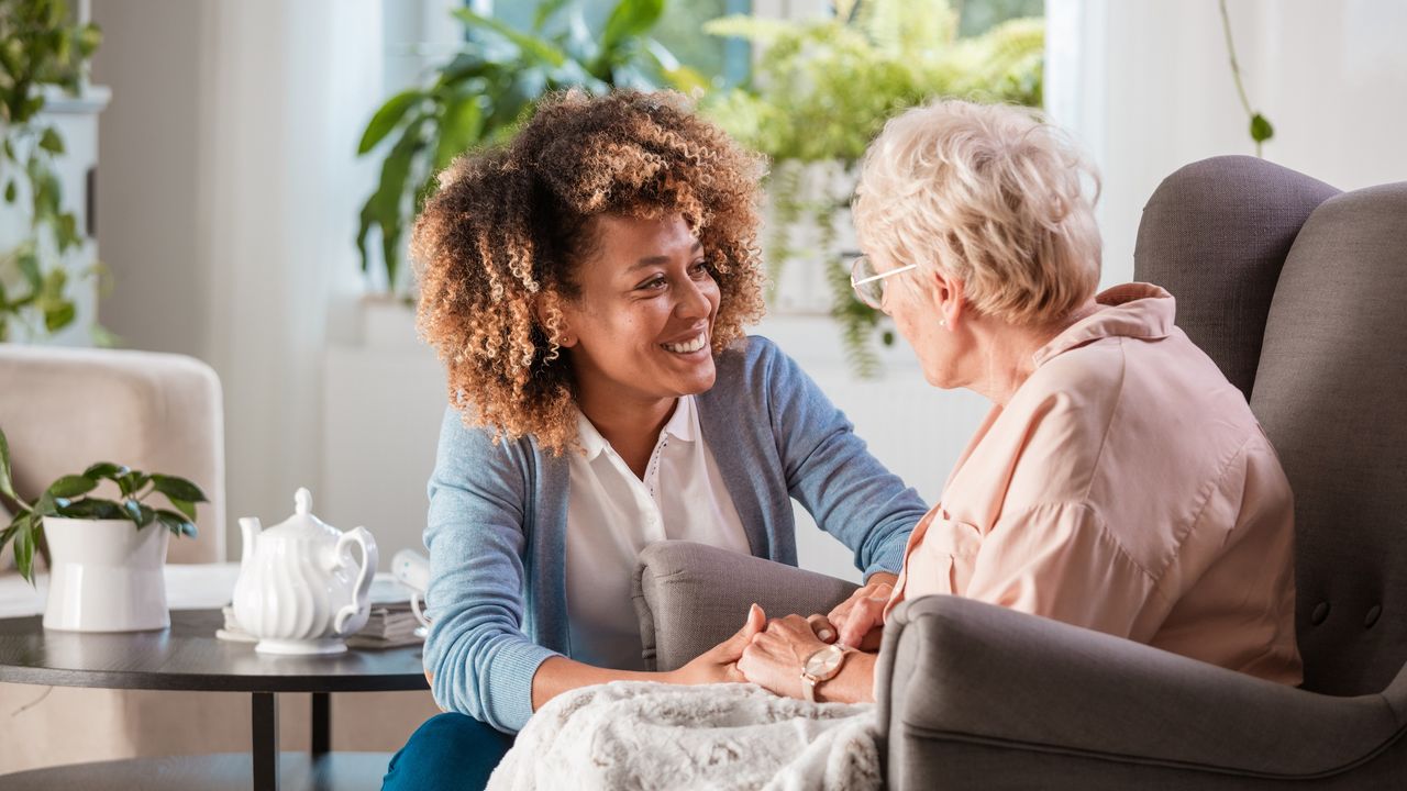 A smiling young woman kneels beside a smiling older woman and holds her hands at a health care facility.