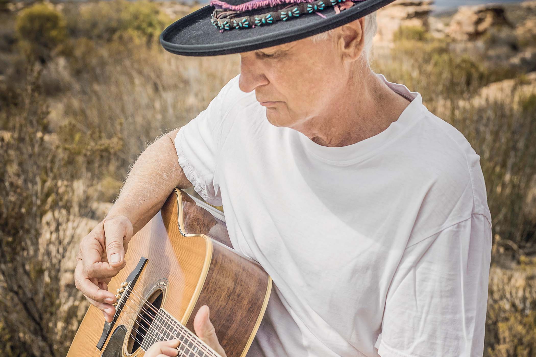 Steve Louw is picture in a wide-brimmed hat with his guitar in the desert.