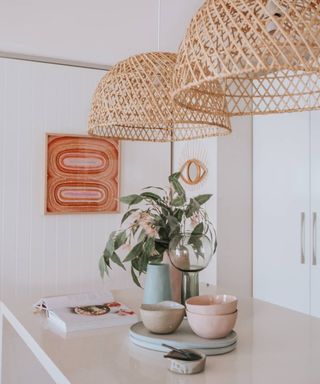 Focal point raffia pendant duo above breakfast counter in white kitchen, with rustic florals in vase, pastel dinnerware, and eye design mirror on wall.