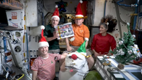 Four astronauts in Santa hats show off their Christmas meal near a Christmas tree on the Interanational Space Station