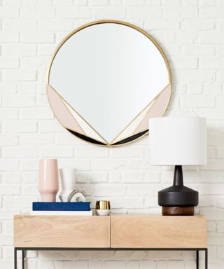 A gold and pink round mirror on a white wall above a wooden console table with books and a lamp on it