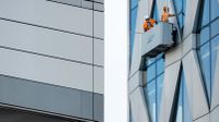 Two workers in orange vests and helmets clean windows from a suspended platform on a modern glass building