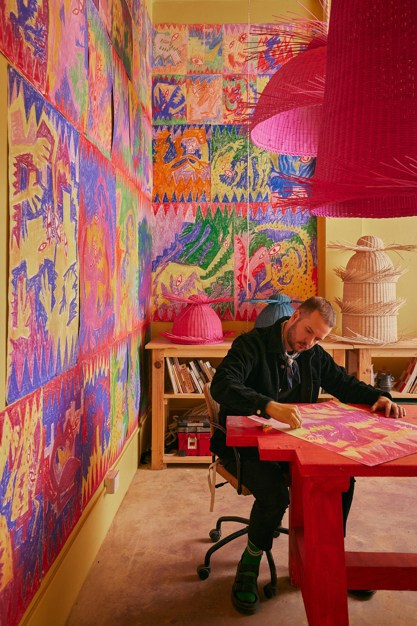 A man in a cargo jacket, with a denim shirt underneath it and matching denim jeans, sits at a red desk working at red, pink, and yellow sketches, in a sketches-filled yellow studio with rattan pieces.