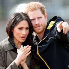 Prince Harry points out something to Meghan Markle, he is wearing a black raincoat with yellow detailing and Meghan is wearing a khaki green jacket