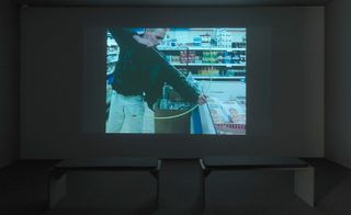 White darkened room, grey floor, two stool seats centre, projection screen on the wall, man in a supermarket, holding a bow aiming an arrow into an open frozen compartment in the middle of a shopping aisle, surrounded by stacked shelves of food items