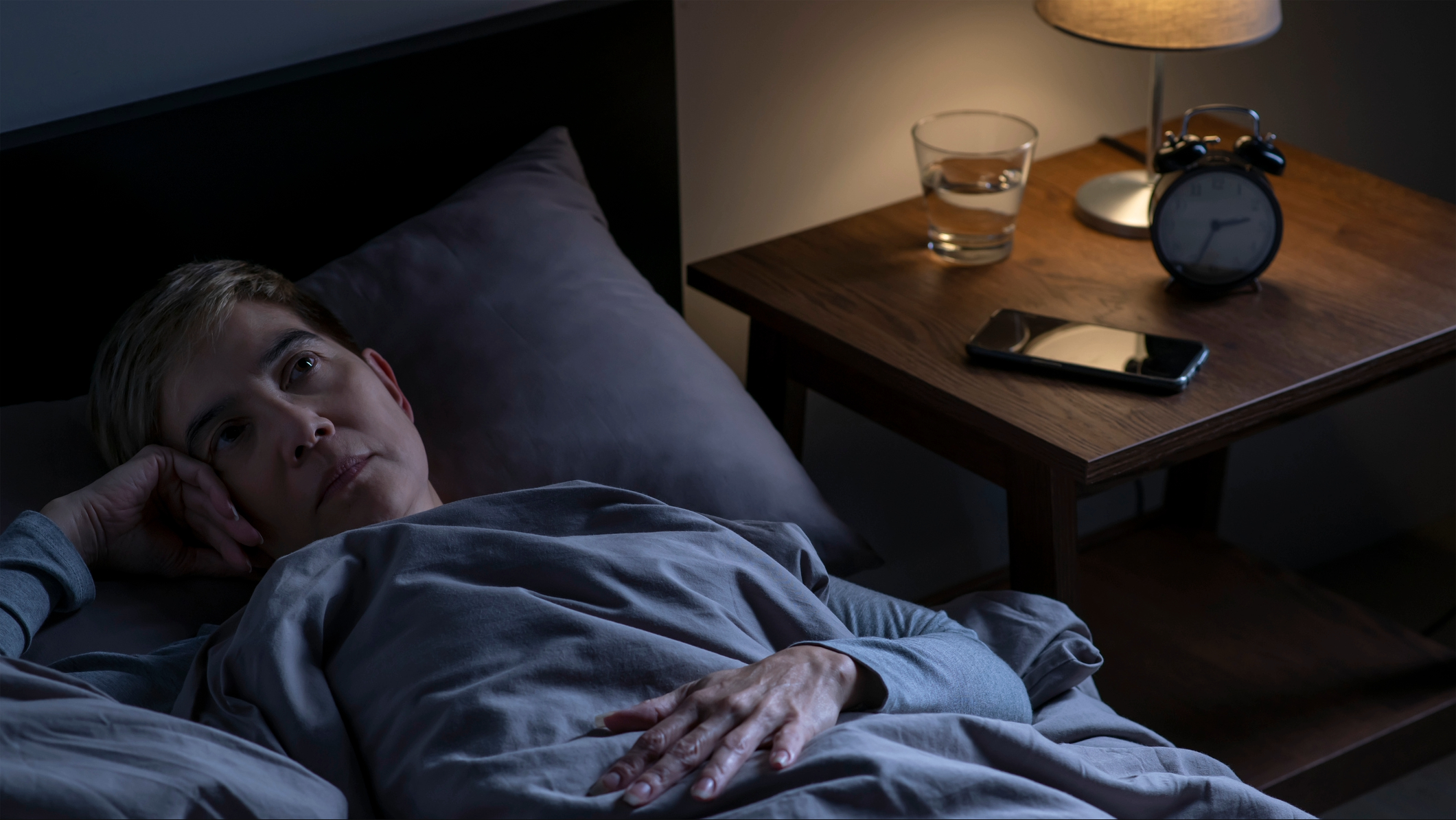 Person lying awake in bed with a lighted lamp next to her with a contemplative expression on her face