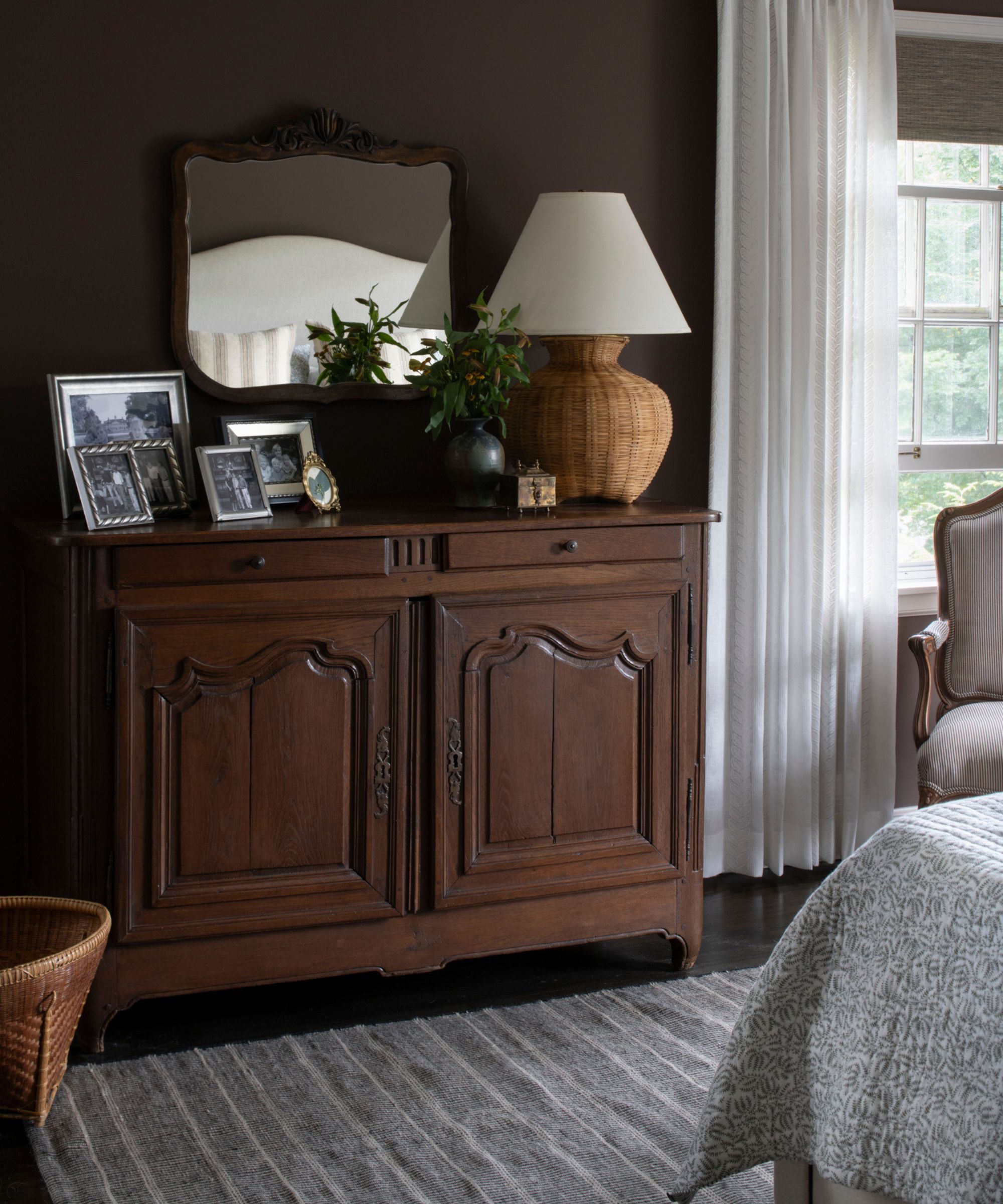 A corner of a bedroom with dark brown walls, a dark wooden vintage dresser with a woven lamp, a wall mirror, and picture frames. A gray rug and a window with sheer white curtains.