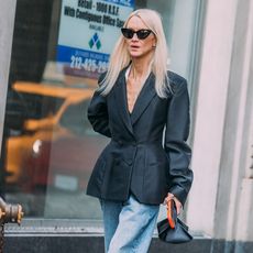 A woman wearing a black blazer, blue jeans, and black cat eye sunglasses