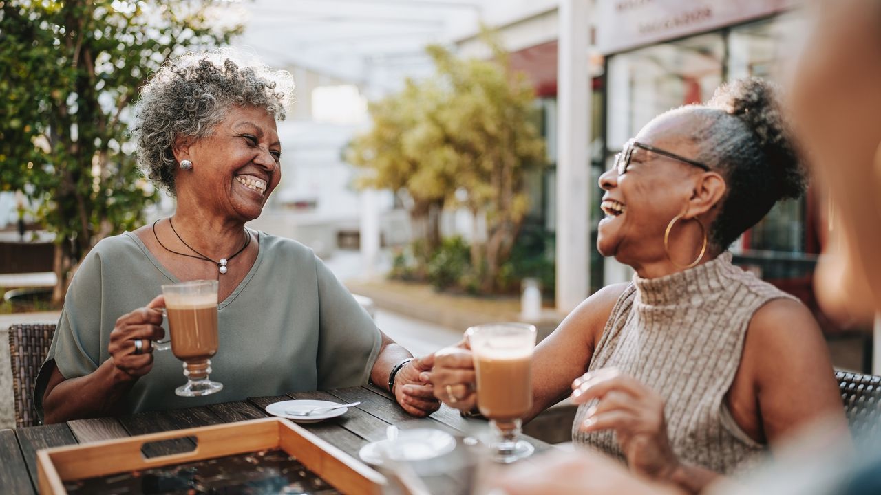 Two older women laugh while drinking coffee at an outdoor cafe.