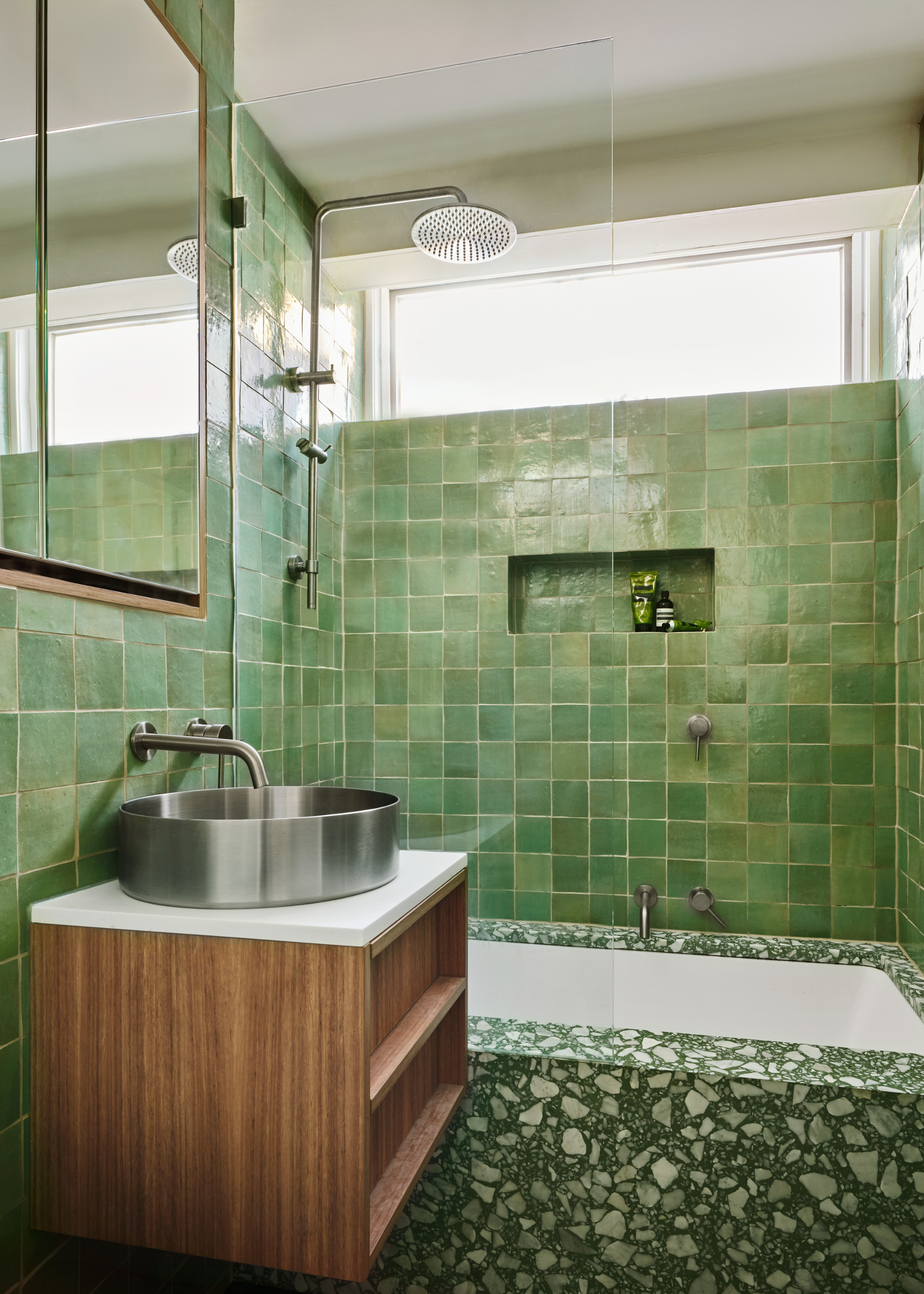 A bathroom with green tiles, a terrazzo bath tub, a wood floating cabinet with a chrome sink, a mirror, and an in-built nook with bath products