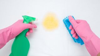 A person wearing pink rubber gloves takes a spray solution and scrubbing brush to a yellow sweat stain on a white mattress