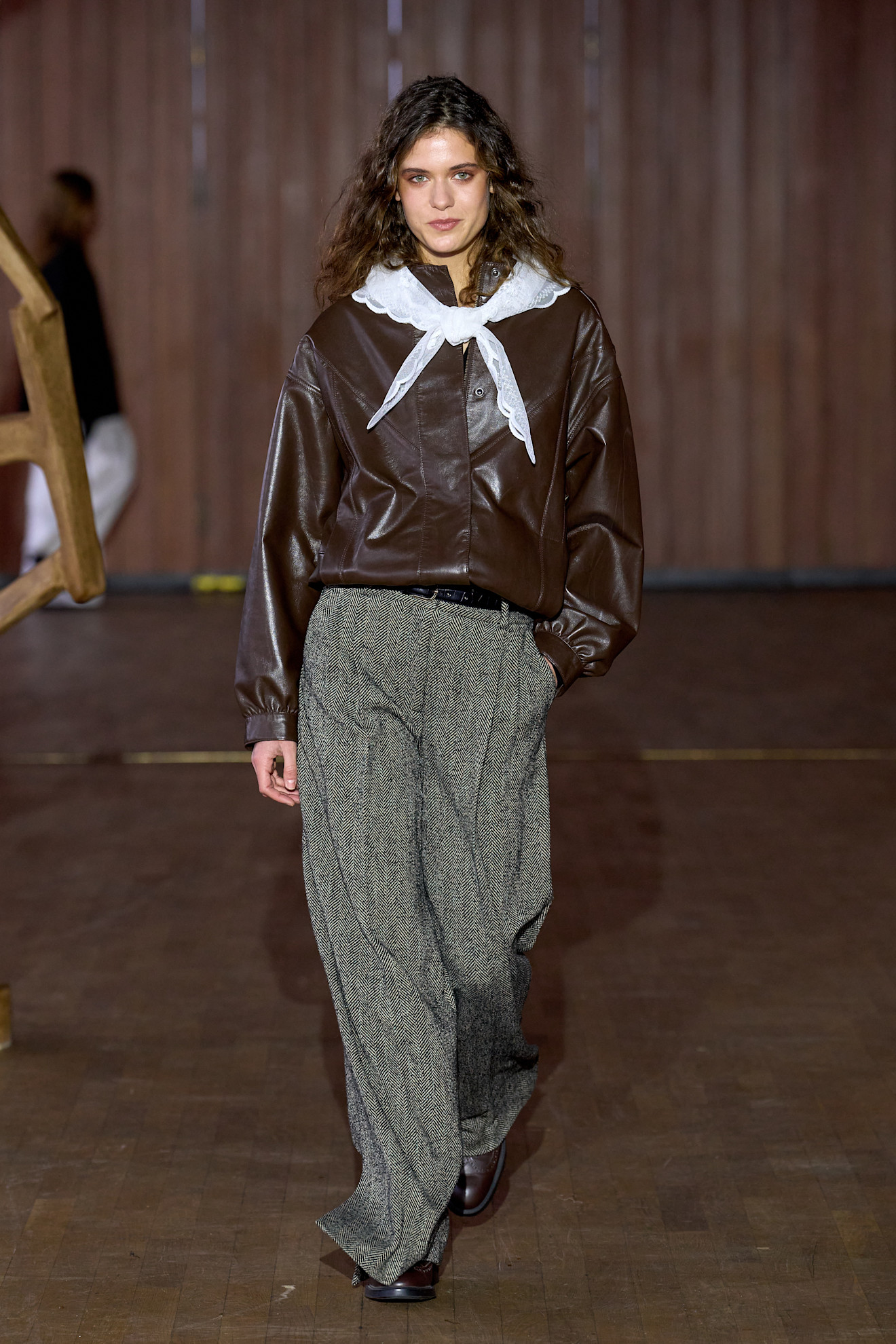 a model at The Garment Fall 2026 show wearing a brown leather jacket, white lace tirangle scarf, and gray pants