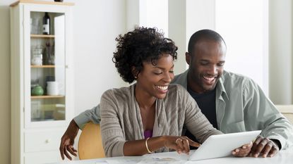 A smiling young couple look at a tablet together while sitting at their dining room table.