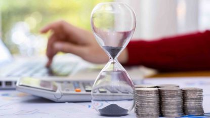 photo of an hourglass next to stacks of coins with a hand in the background working on a calculator