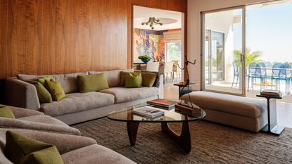 Image of a room with wood-panelled walls and glass sliding doors to a sunny yard. There is a gray textured sofa, textured accent chairs, and a textured rug. 