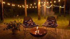 A fire pit on a deck surrounded by two comfortable chairs at night, with the garden lit up