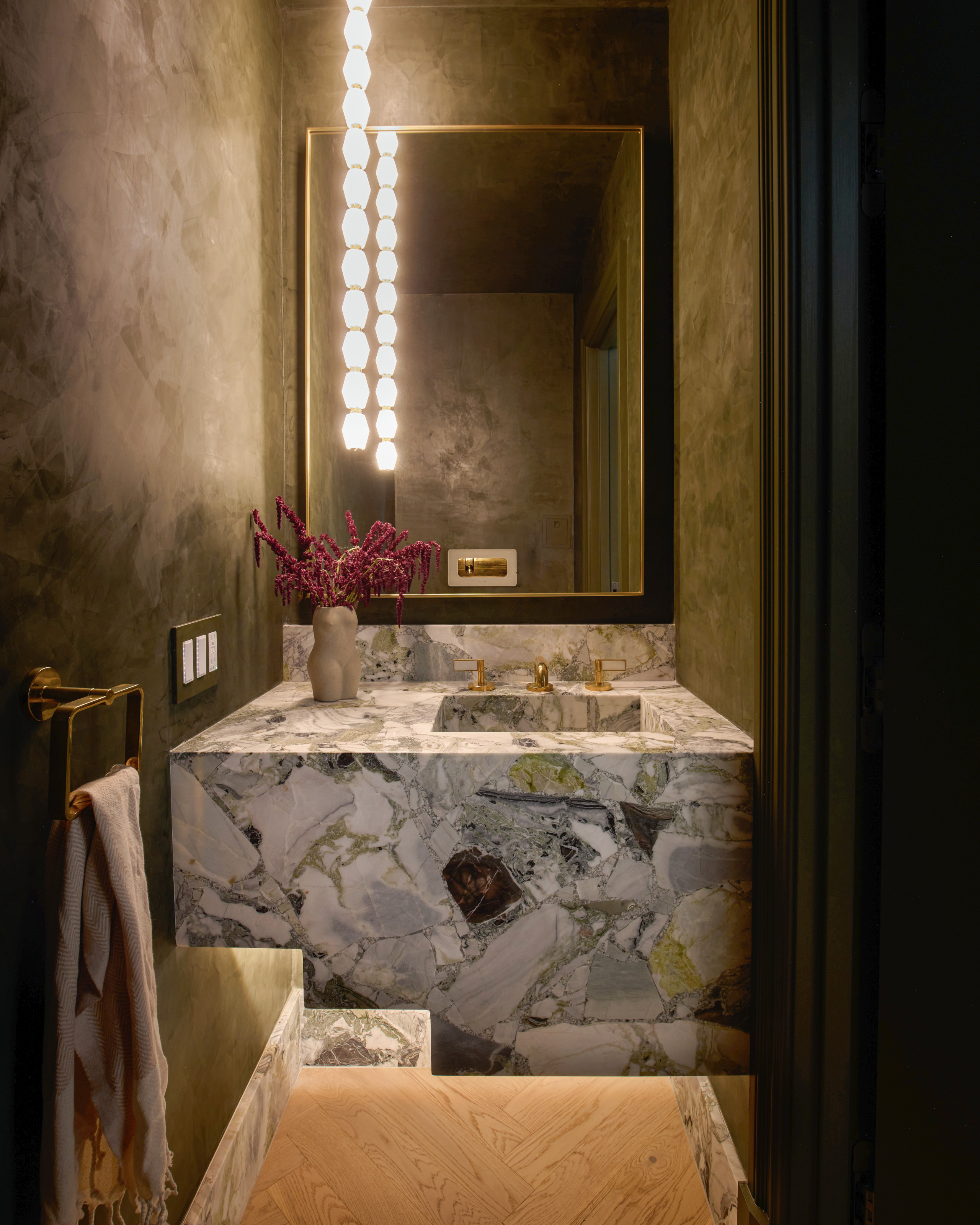 Powder room with green textured walls, green and white stone vanity, rectangular gold mirror and pendant light