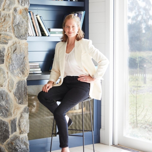 Image of a woman in a white blazer and black trousers sitting on a stool in front of a blue bookscase.