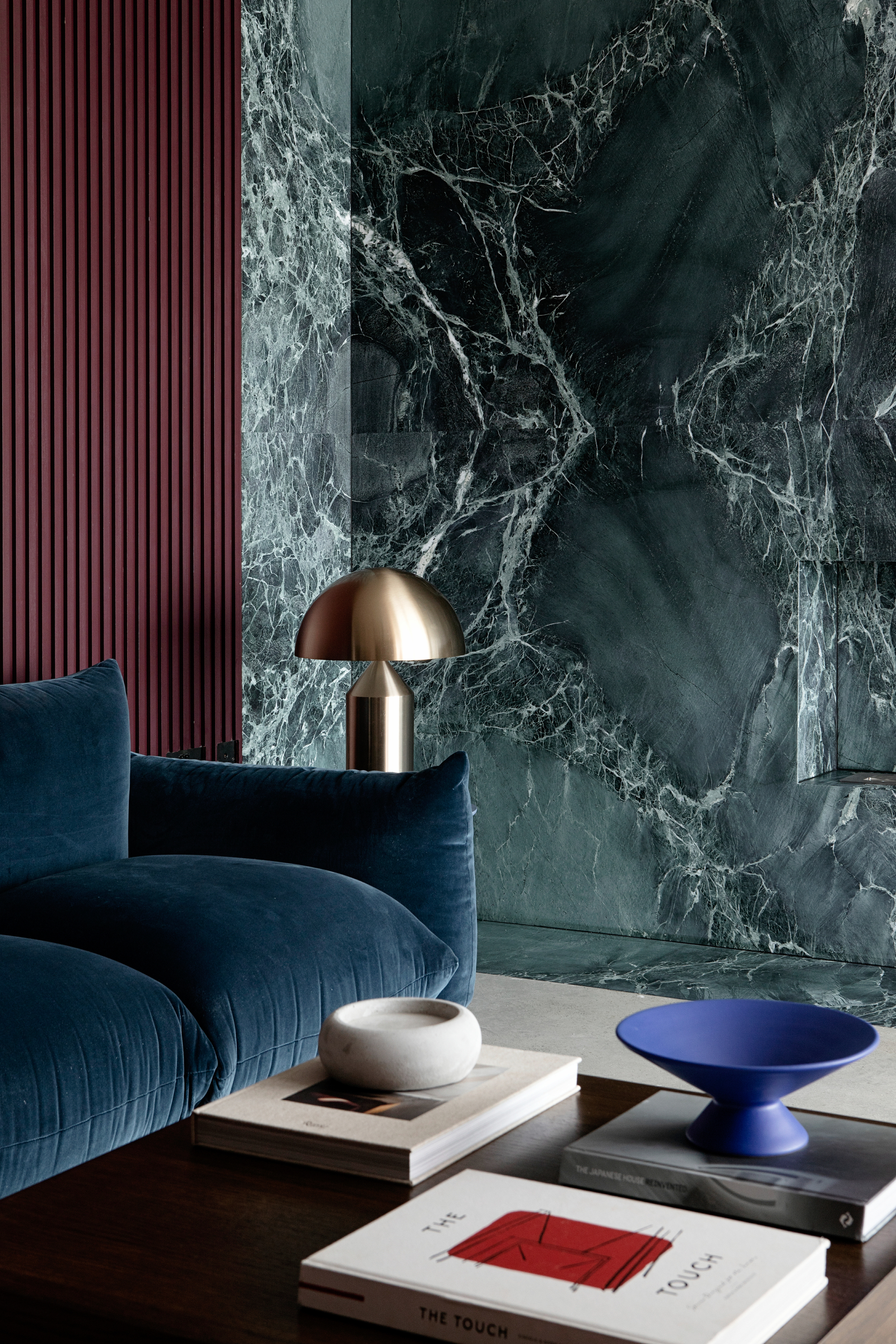 Image of the corner of a living room where the walls are a teal and white marble. There is a dark teal sofa and a large black coffee table with multiple white books and a cobalt blue decorative bowl. There is also a chrome mushroom lamp.