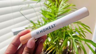 A person holding a white mascara tube with a light coral pink end and the company name Honest printed on the mascara tube in black, for the Honest Beauty Extreme Length + Lash Primer review.