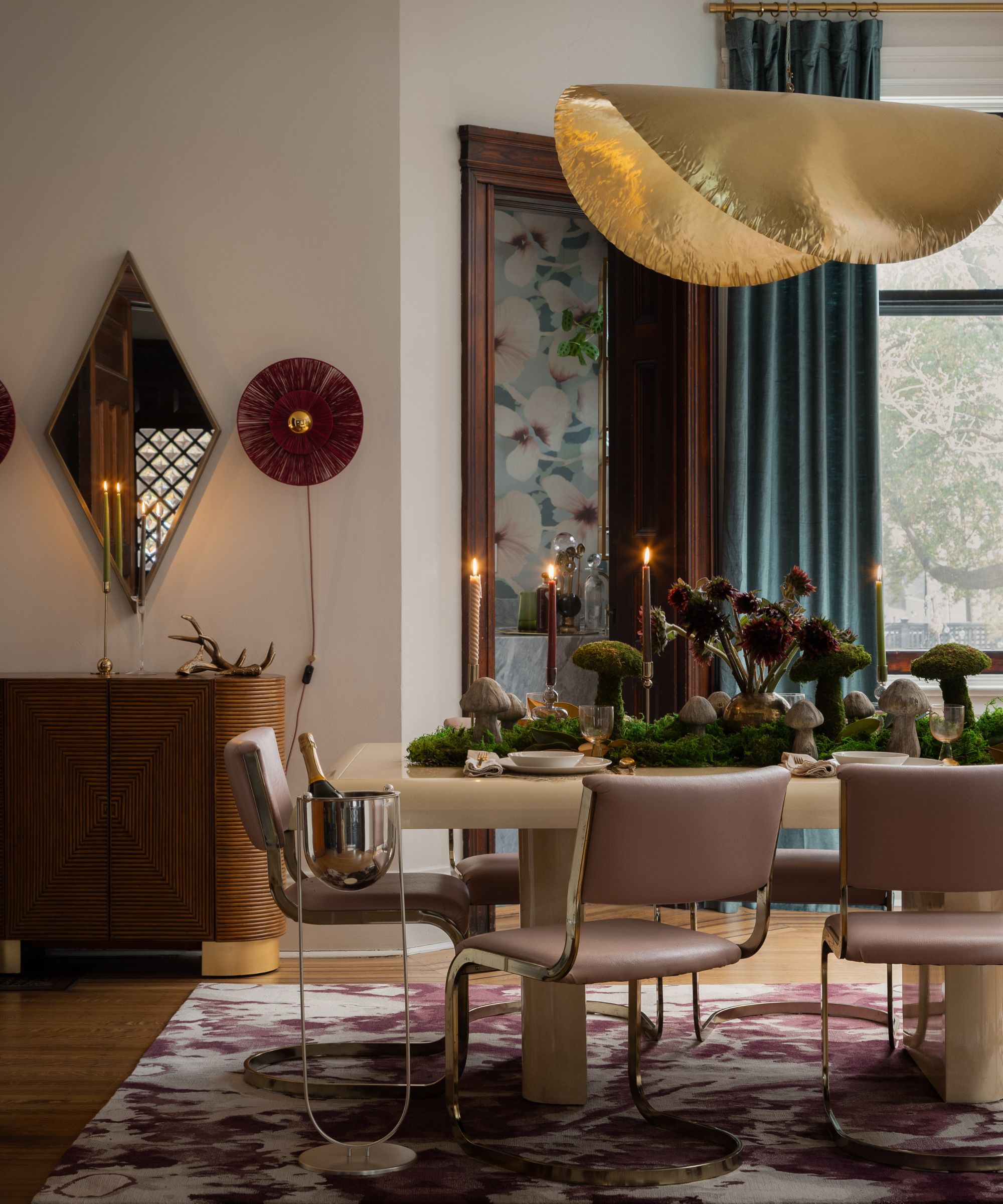 Dining room with a gold, wavy pendant light, pink leather chairs, a wood cabinet, and a table decorated with moss and mushrooms