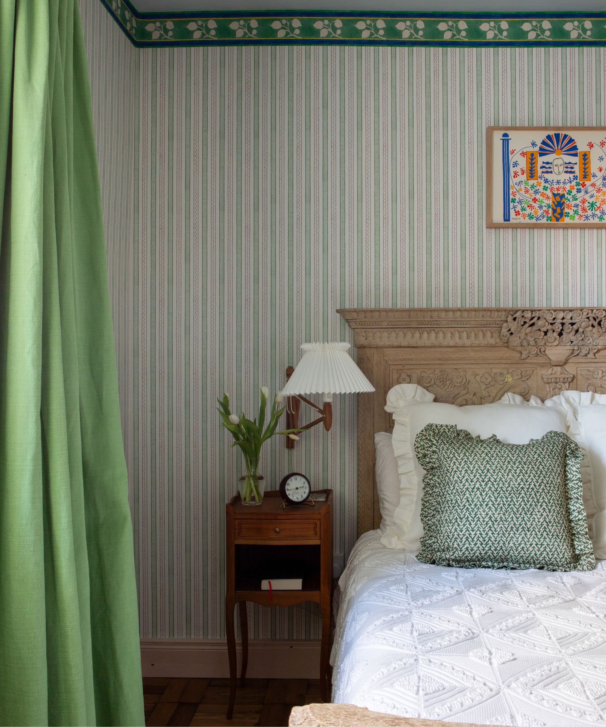 Bedroom with striped green wallpaper, carved wooden headboard with white bedding and green frilly cushion, a wooden side table with a pleated sconce above, and green long drapes covering the window