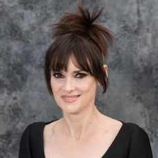 a white woman with dark hair wearing a spiky ponytail and bangs in front of a gray background