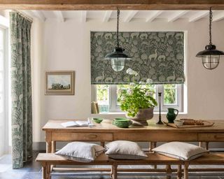 A dining room with a large wooden table with matching benches either side of it, softened with cushions. Two large black rustic pendant lights hanging over the table. In the background, a large window is covered by a green patterned roman blind.