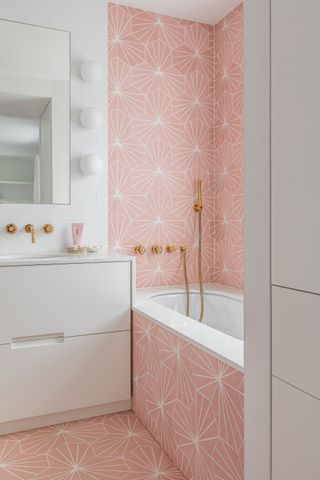 white bathroom with pink patterned floor and wall tiles, gold fixtures and fittings, white chest of drawers, white wall tiles, mirror, globe lights