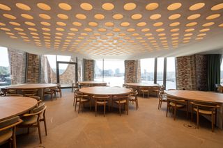Fjordenhus, by Olafur Eliasson and Studio Olafur Eliasson in Vejle, Denmark. A dining area with round tables, chairs, large windows and round roof lights overlooking a body of water.