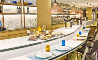 A restaurant with a long white marble counter with a variety of cheese on it and chairs in front of the counter and shelving behind.