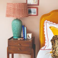 A pink-painted bedroom with a mustard yellow velvet headboard, a patterned table lamp, a scallop-trim bed linen and a leopard-print cushion