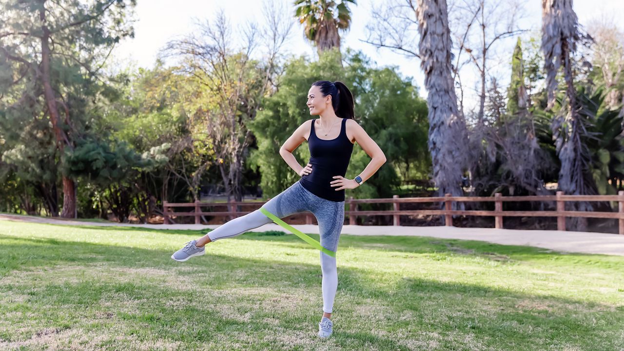 A woman performs a resistance band workout in a park, wearing leggings, sneakers and a vest. She is standing on her left leg with the band looped around her thighs, keeping her right leg straight and pushing it out to the side. Her hands are on her hips and she wears a ponytail and fitness tracker.