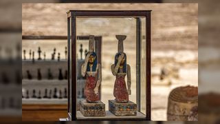 Statuettes depicting the Egyptian goddesses Isis (left) and Nephthys (right). Both statuettes have gold faces, dark blue headpieces, necklaces, white tops, and long red skirts. They are kneeling and carrying pottery on their heads.