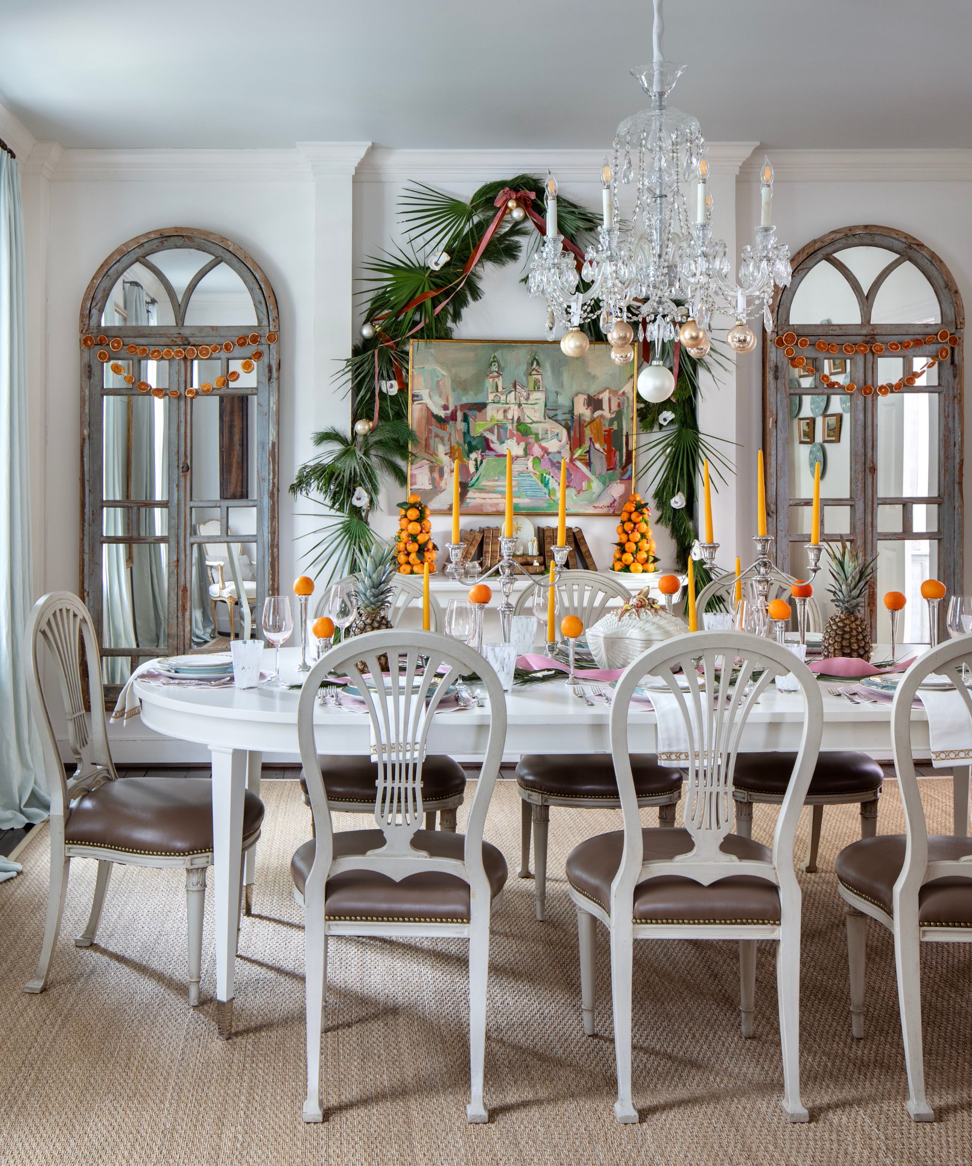 Grand dining room with a long white dining table decorated with taper orange candles, clementines and silverware. A large fireplace sits in the centre, decorated with a garland.