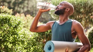 a photo of a man carrying a yoga mat in one hand and drinking from a water bottle in his other hand