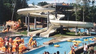 A packed water park on a sunny day with hundreds of smiling visitors jumping in pools and down water slides.