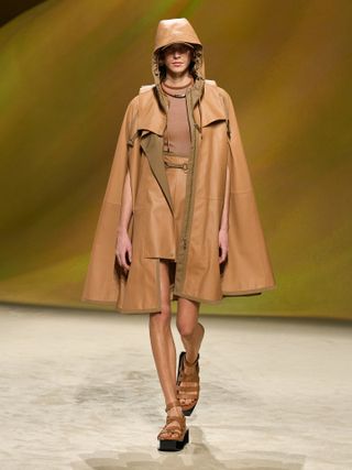 A model wearing a light brown hooded leather knee high coat and shorts and leather sandals.