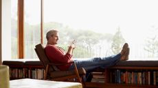 Full length side view of thoughtful mature man relaxing on armchair by bookshelves