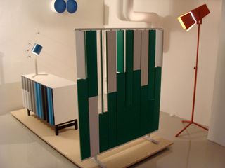 A furniture collection featuring a green and white room divider, white cabinet with horizontal lines (grey, blue and white) with a blue table lamp placed on it. On the right corner is a red standing lamp,