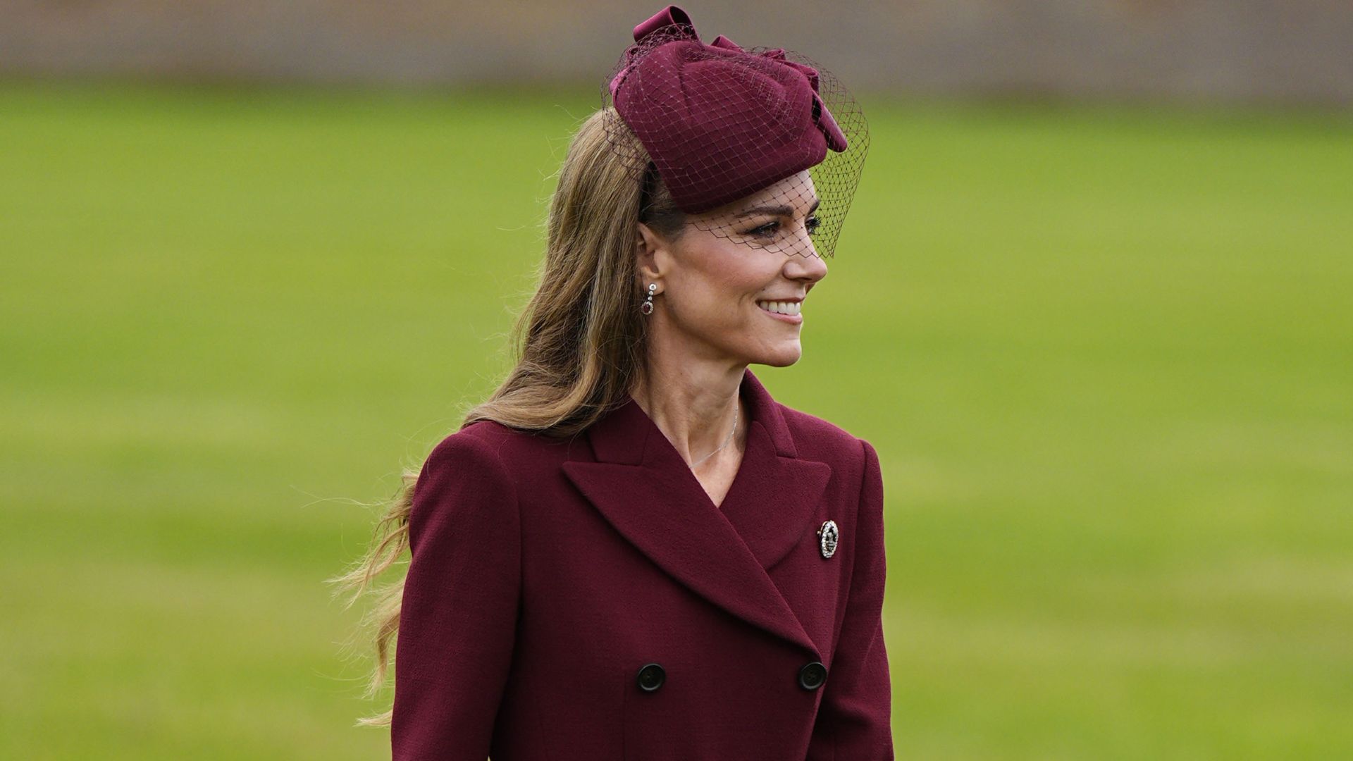 Kate Middleton walks outside on grass wearing a burgundy coat with chocolate brown heels. She carries a burgundy Chanel bag.