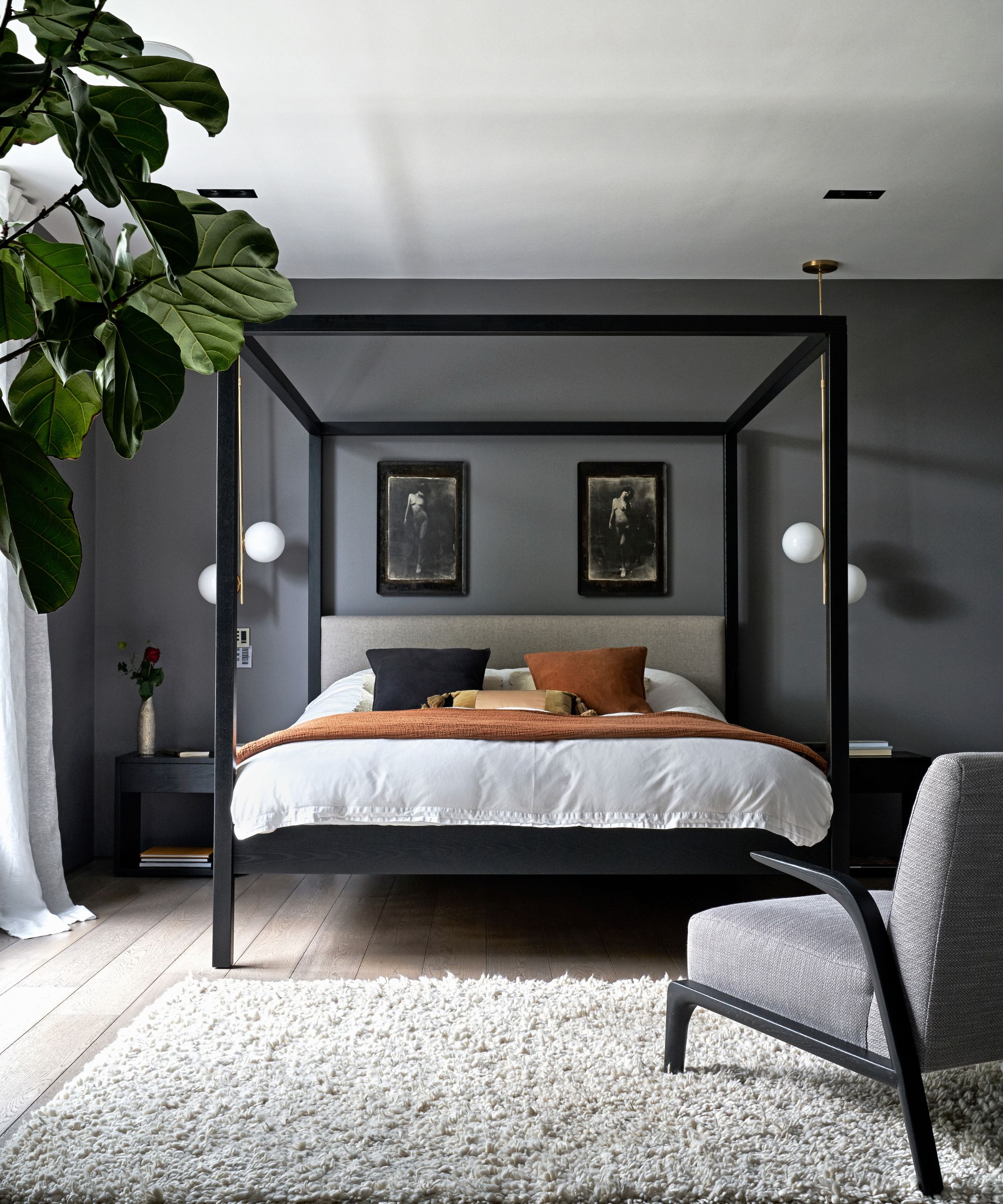 Gray bedroom with a white rug, black four-poster bed with white bedding, cushions, a gray upholstered chair, and a large houseplant to the left.